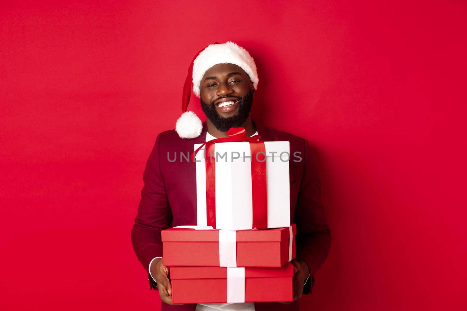 Christmas, New Year and shopping concept. Happy Black man in santa hat and blazer holding xmas presents, bring gifts and smiling, standing against red background.