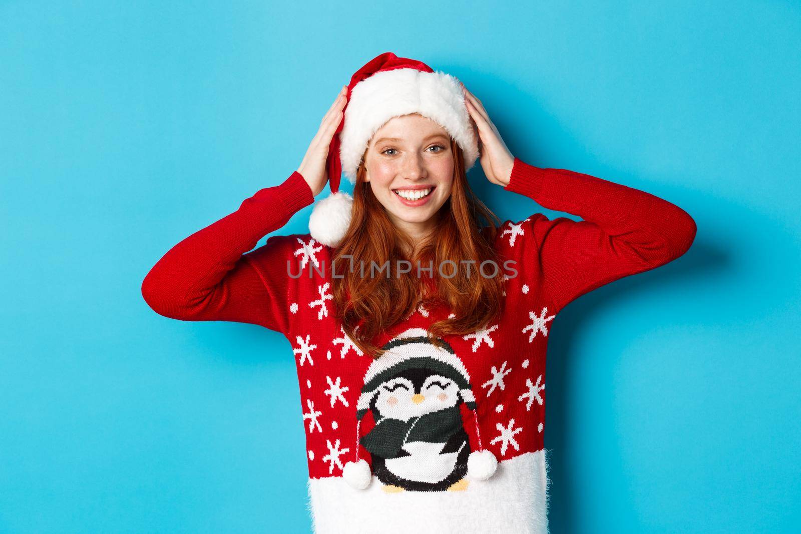 Happy holidays and Christmas concept. Pretty redhead girl in xmas sweater, put on santa hat and smiling, celebrating New Year, blue background by Benzoix