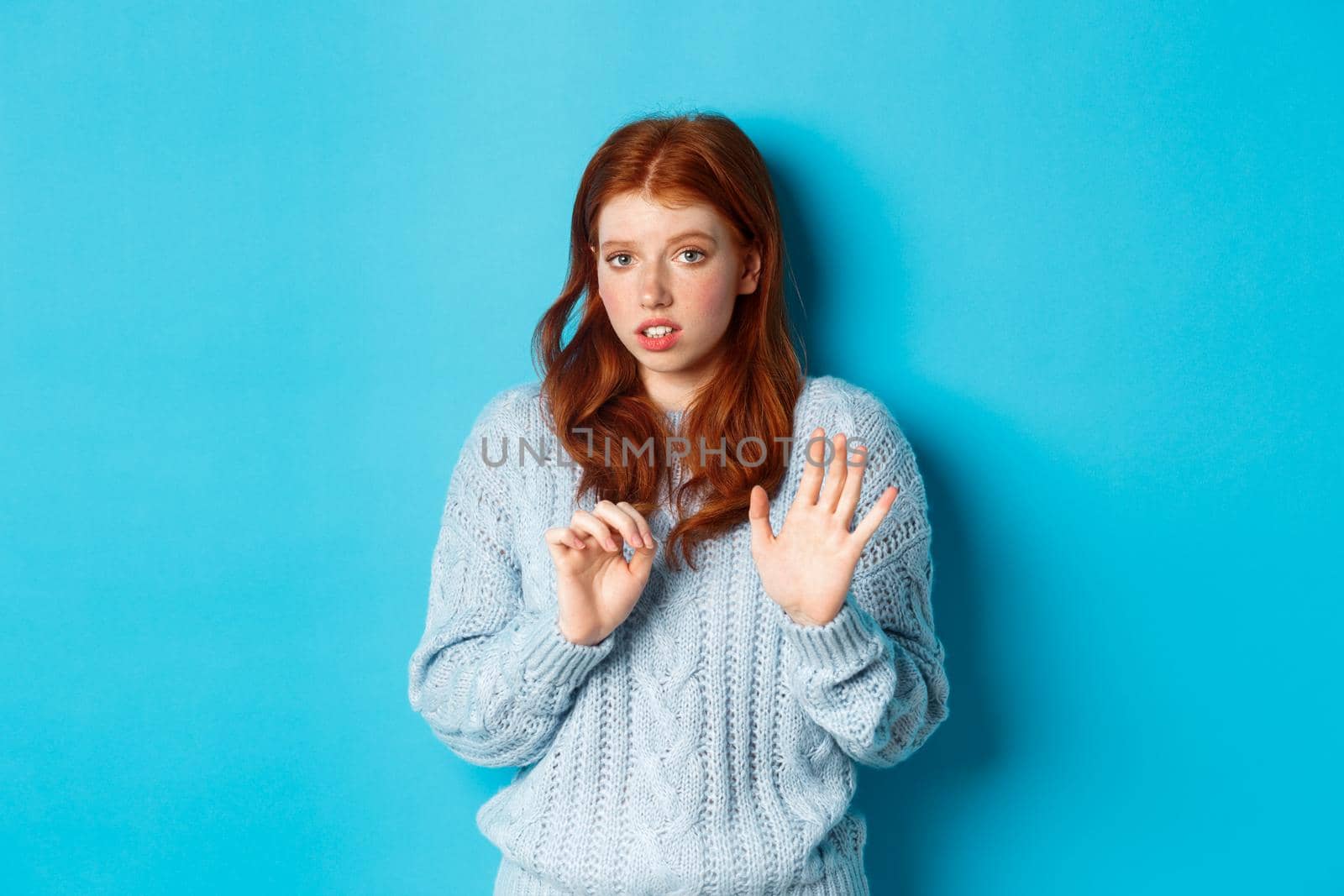 Worried redhead girl refusing or declining an offer, shaking hands and looking anxiously at camera, rejecting something unpleasant, standing over blue background.
