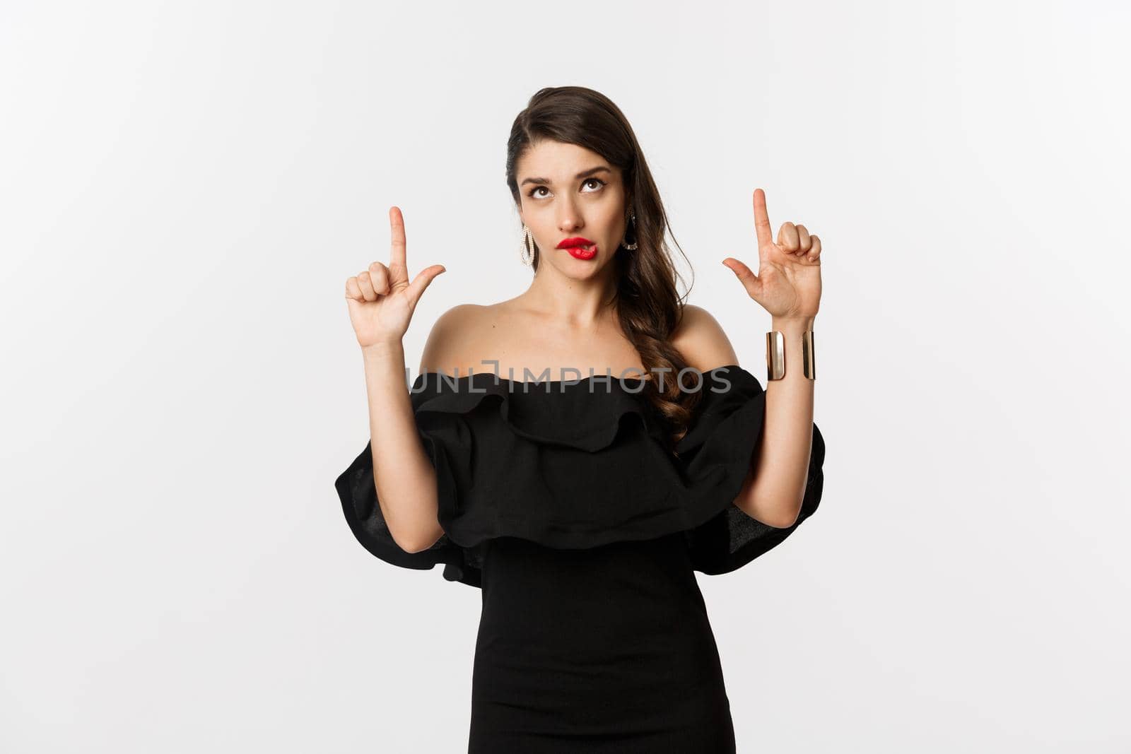 Fashion and beauty. Thoughtful attractive woman in black dress staring and pointing up, thinking with tempted look, white background.