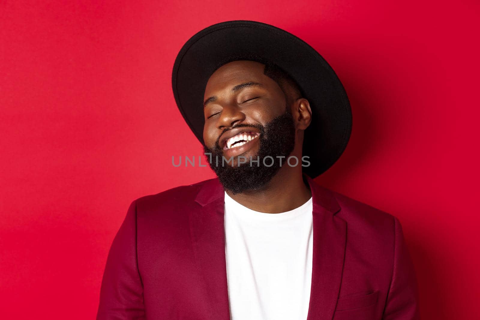 Close-up of handsome Black man with long beard laughing and having fun, looking carefree, standing over red background.
