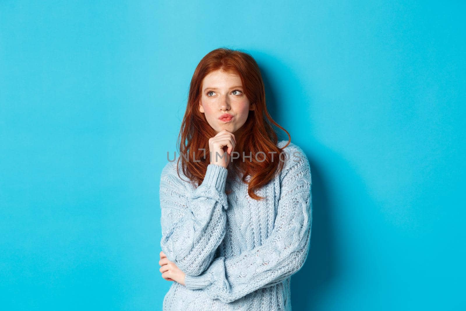 Pretty teenage girl with red hair thinking, looking upper left corner thoughtful, pondering something, standing over blue background.