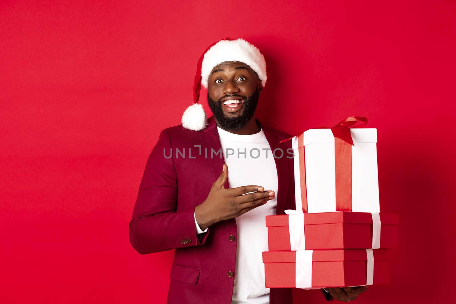 Christmas, New Year and shopping concept. Cheerful Black man secret santa holding xmas presents and smiling excited, bring gifts, standing against red background by Benzoix