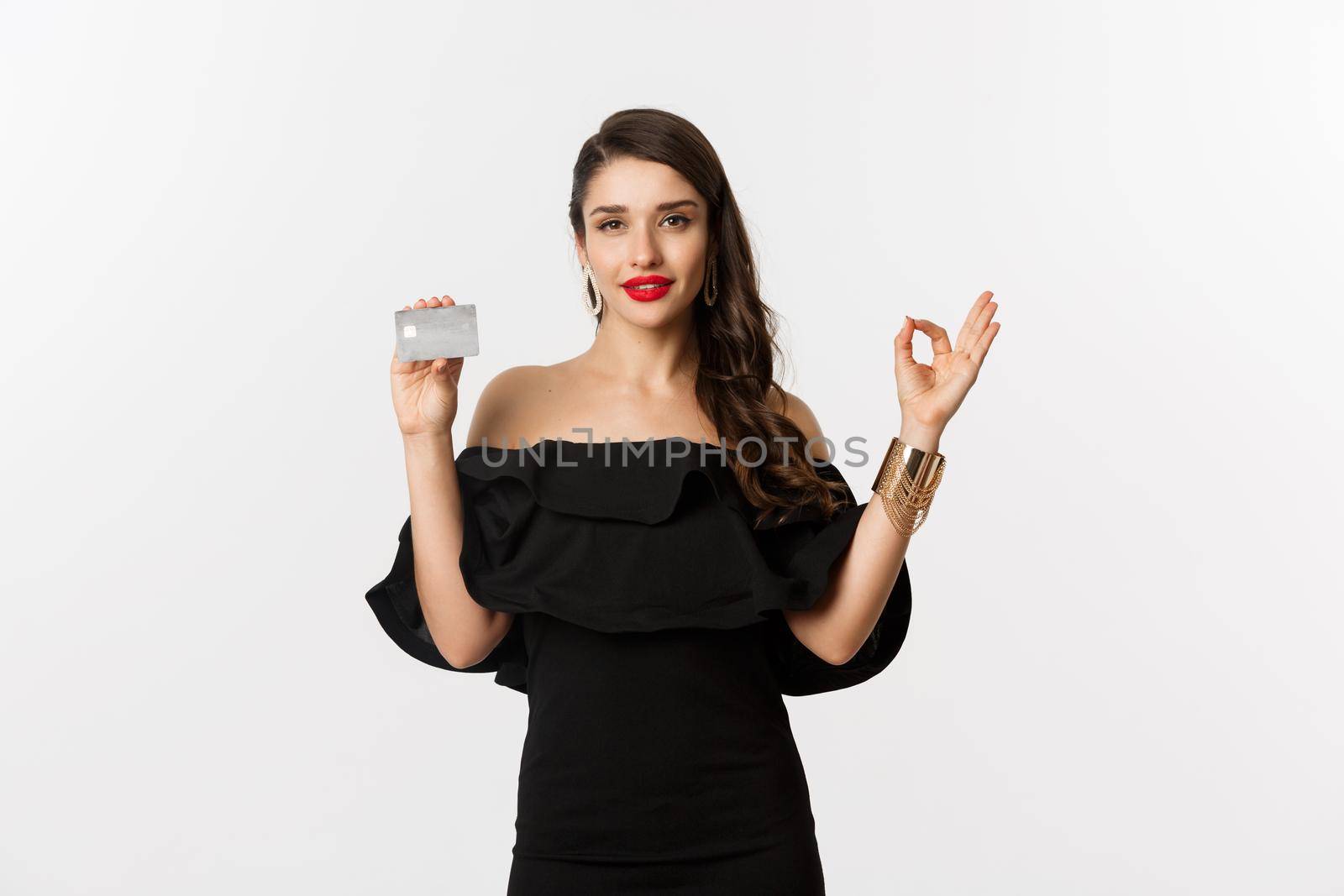 Beauty and shopping concept. Gorgeous woman in luxury jewelry and black dress, showing okay sign and credit card, standing over white background.