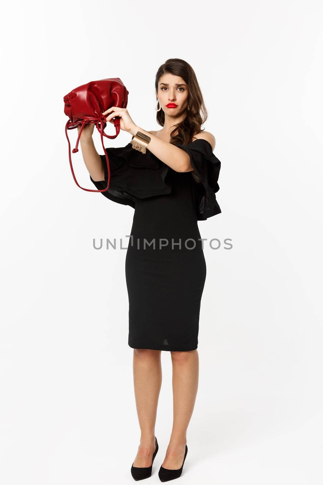 Beauty and fashion concept. Full length of sad young woman in black dress and high heels showing empty purse, sulking disappointed, standing over white background by Benzoix