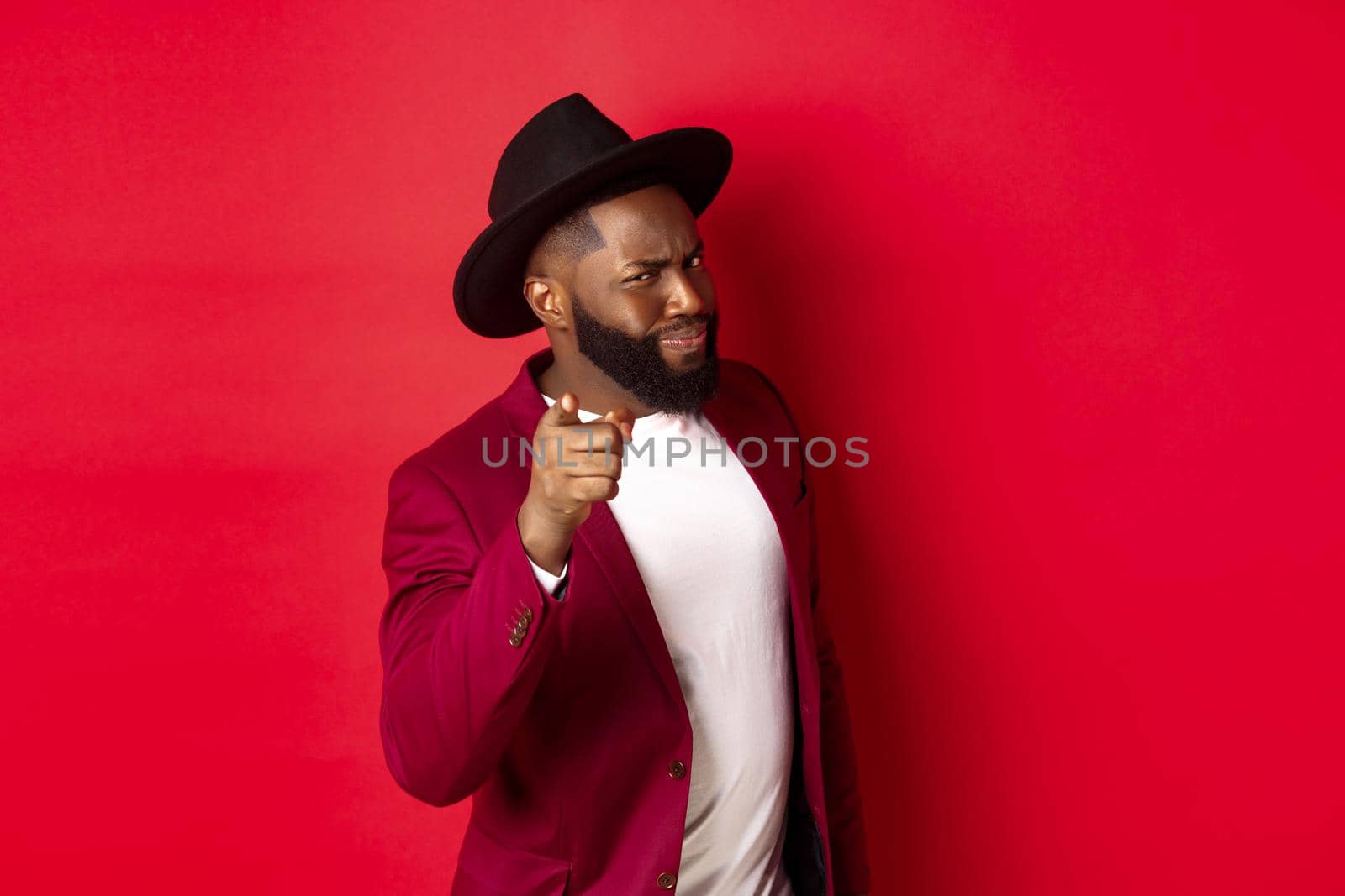 Suspicious Black man pointing finger at camera and squinting, accuse you, give warning, standing against red background.