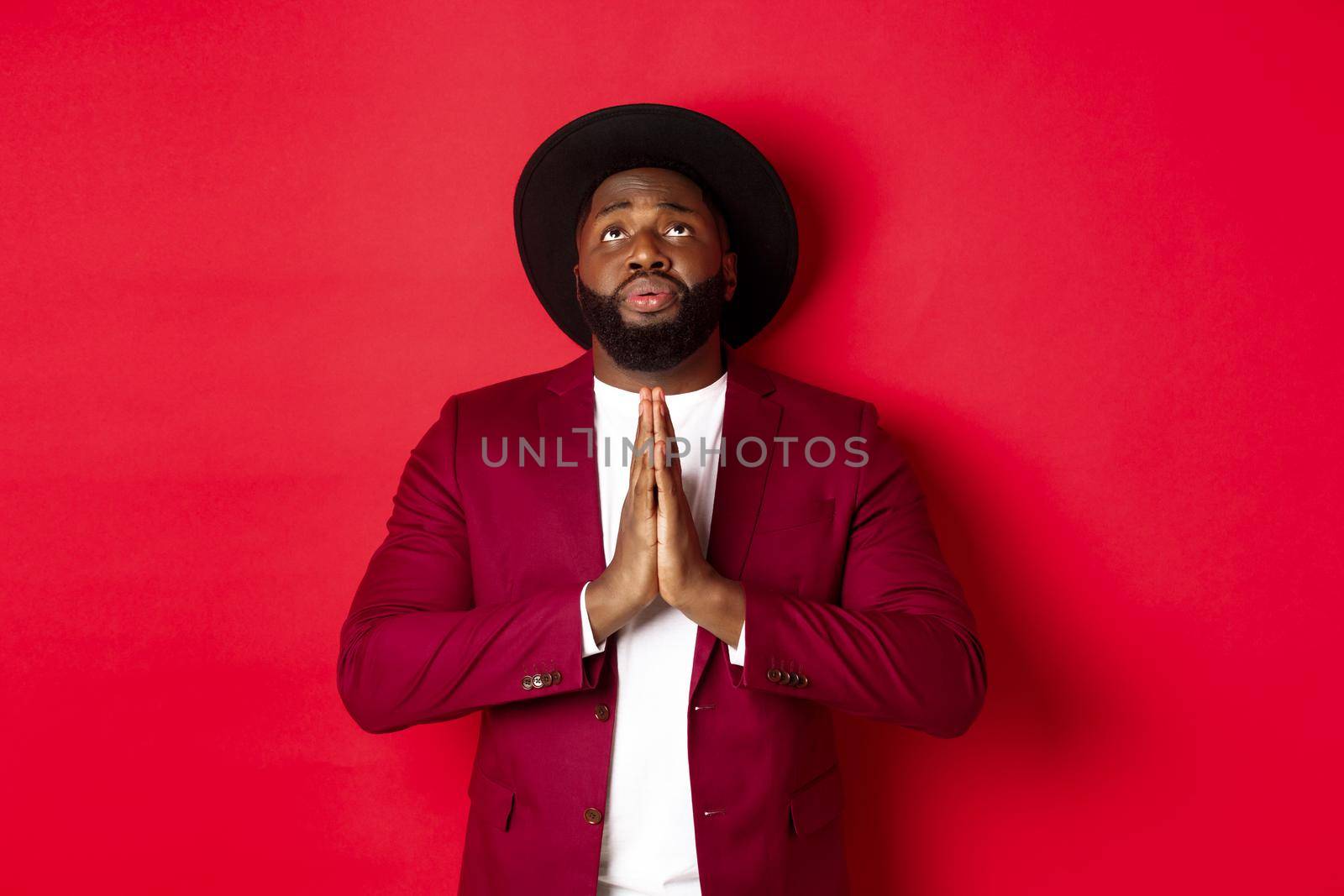 Hopeful and sad Black man talking to God, holding hands in pray and looking up, pleading, begging for help, standing against red background by Benzoix