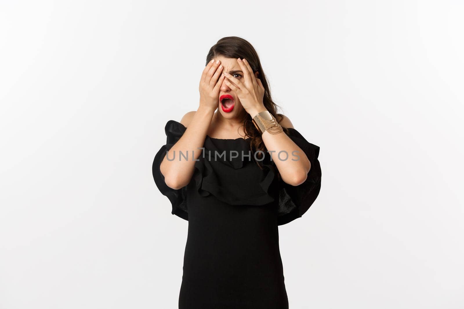 Fashion and beauty. Shocked young woman in black dress covering eyes, peeking through fingers at something embarrassing, cringe, standing over white background by Benzoix