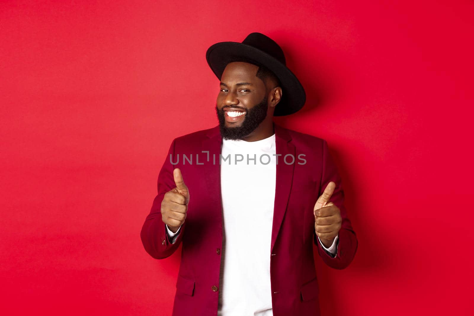 Cheerful Black man having fun on party, showing thumbs up in approval, smiling and liking something, standing against red background.