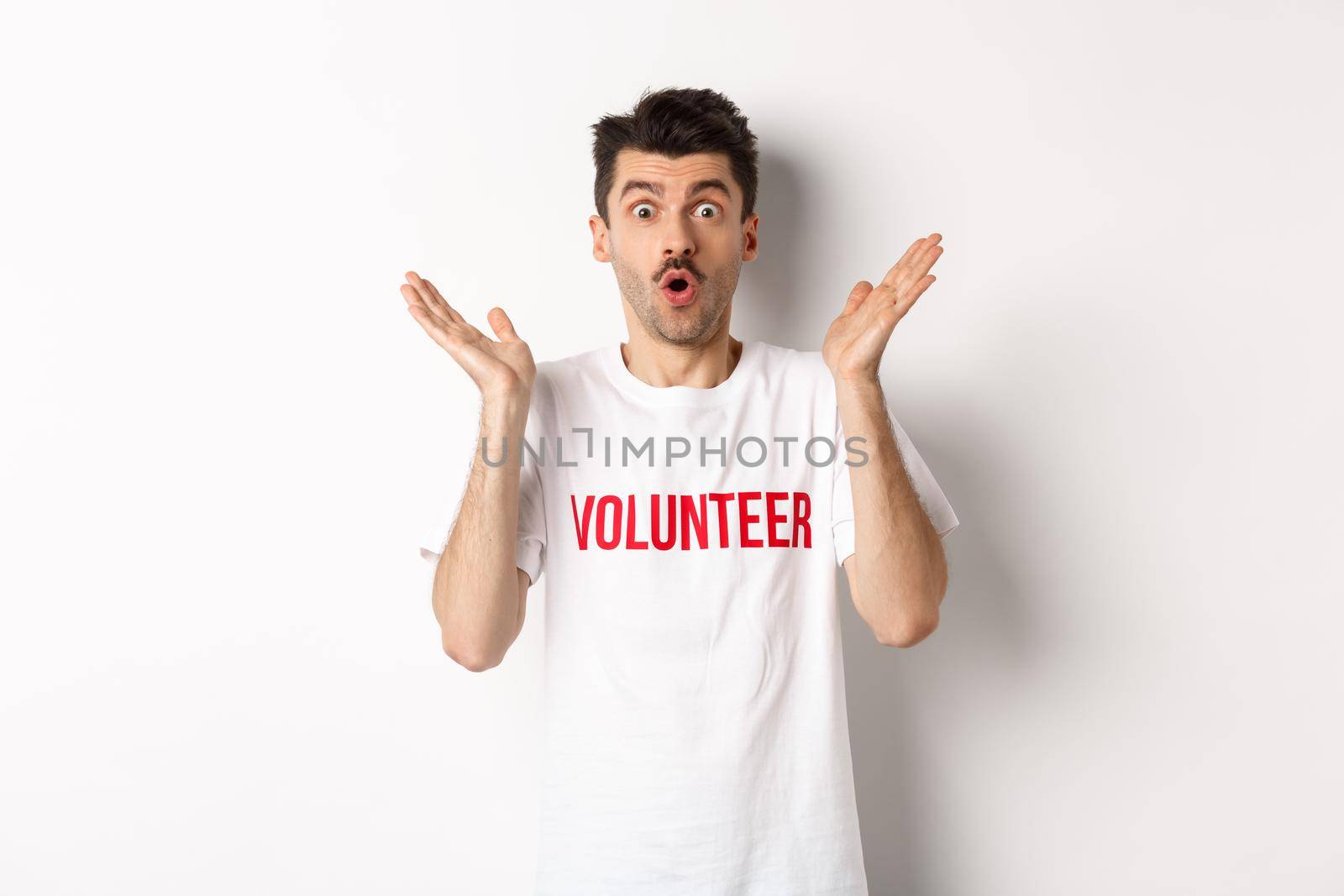 Amazed male volunteer in white t-shirt hear great news, clap hands and staring at camera surprised, posing over studio background.