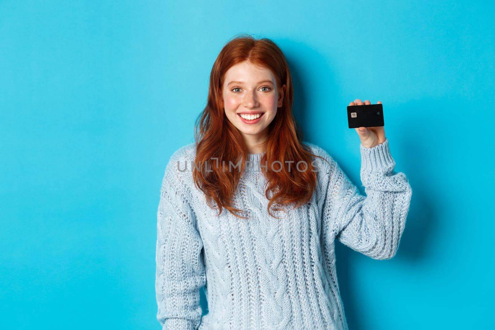 Cute redhead girl in sweater showing credit card, smiling at camera, standing over blue background by Benzoix