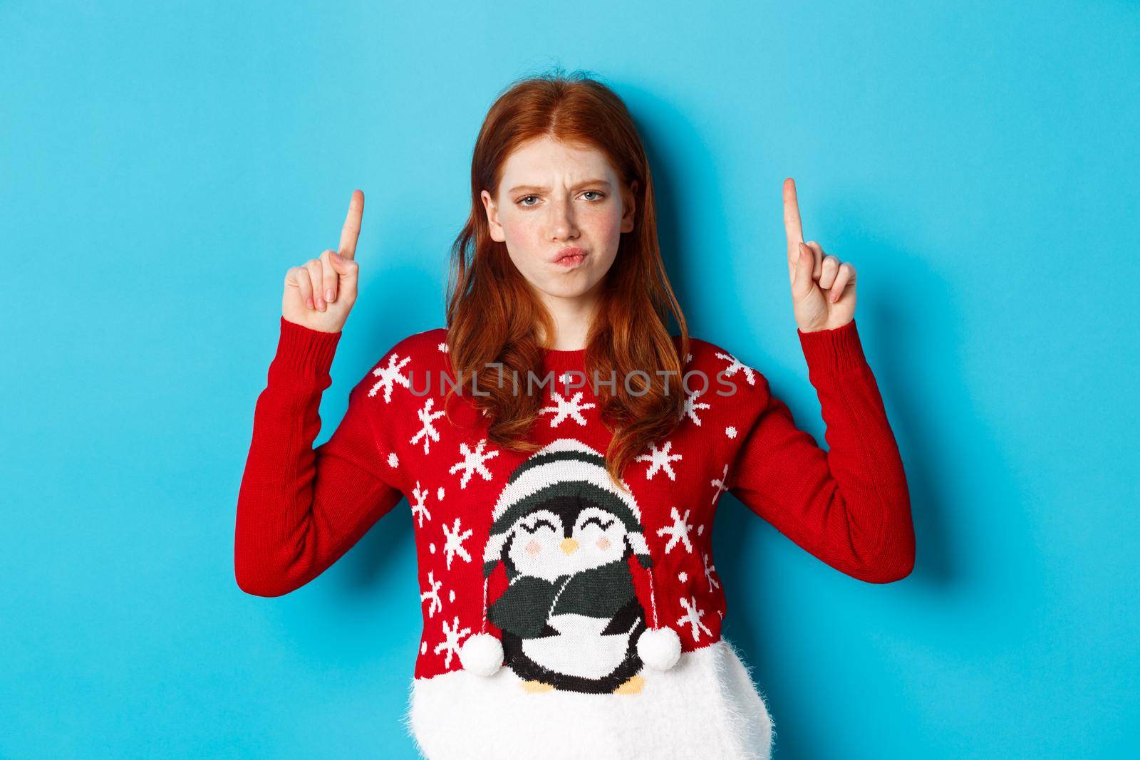 Winter holidays and celebration concept. Perplexed redhead girl in xmas sweater, frowning doubtful and pointing fingers up, showing advertisement, blue background.