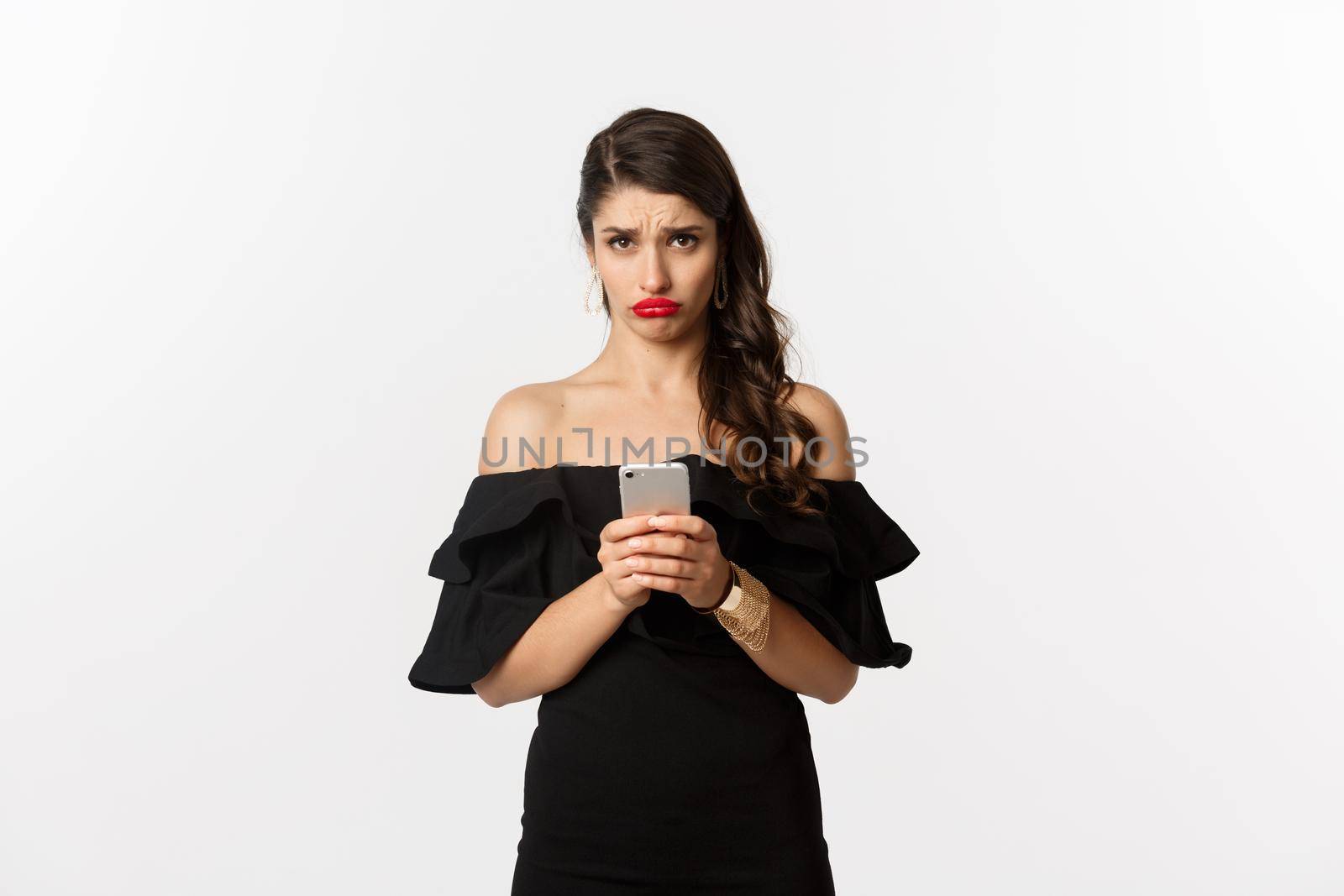 Sad and gloomy woman in black dress, sulking upset, using mobile phone and feeling disappointed, standing over white background.