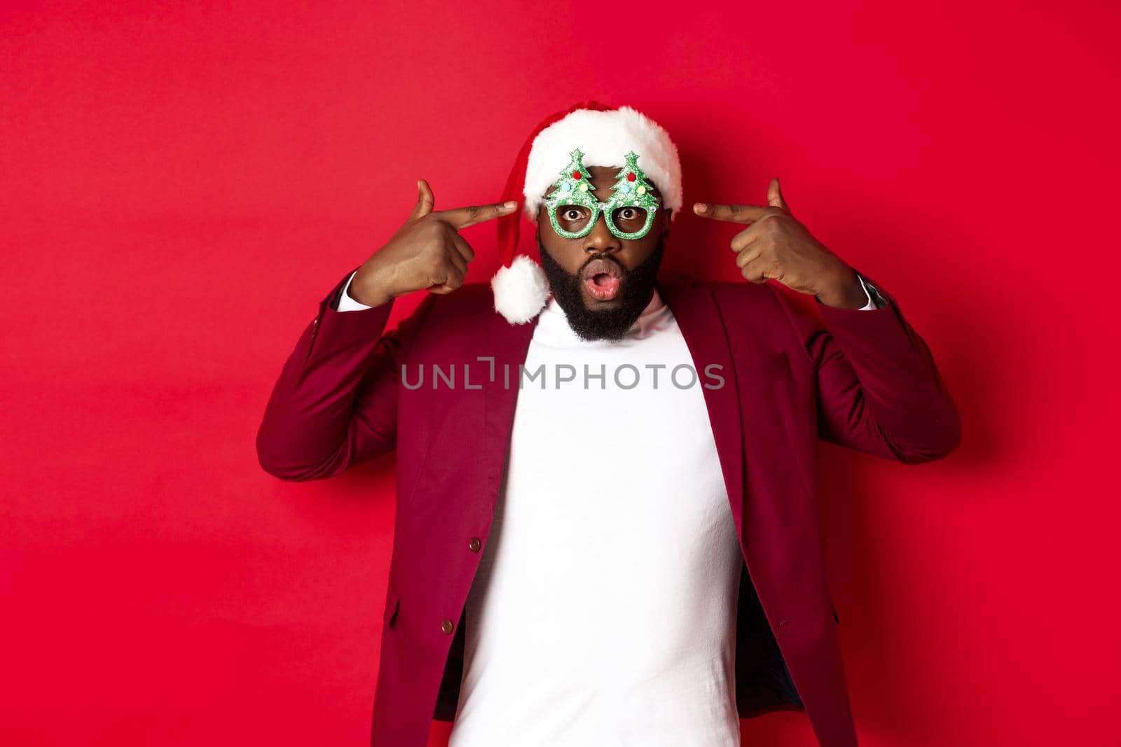 Merry Christmas. Cheerful Black man wearing funny party glasses and santa hat, smiling joyful, celebrating winter holidays, standing over red background by Benzoix