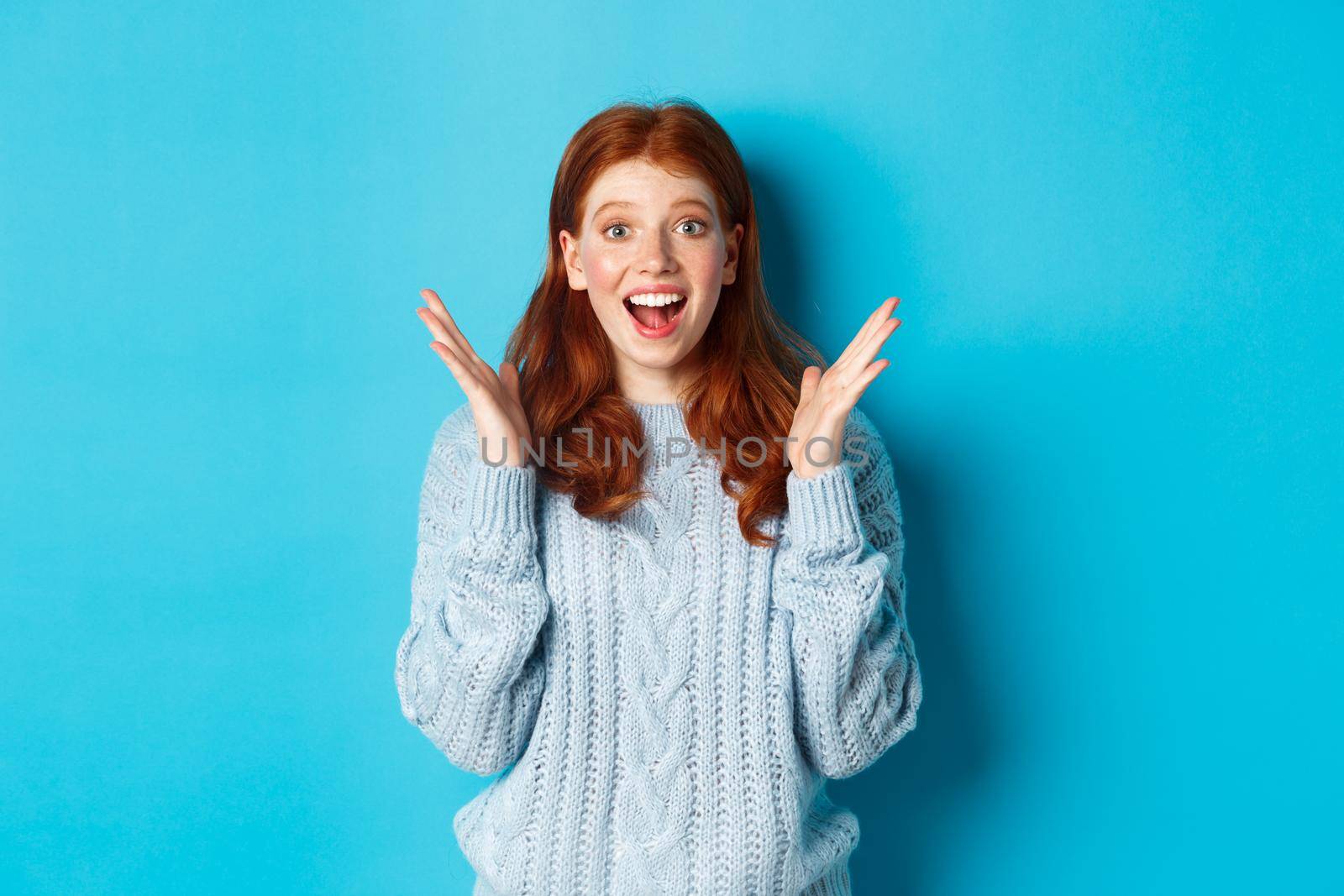 Surprised and happy redhead girl clap hands and staring at camera, smiling amazed, standing against blue background by Benzoix