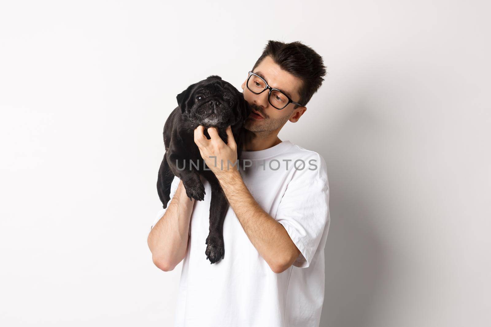 Handsome young man pet his cute black dog, scratching pug while holding animal on shoulder, standing over white background.