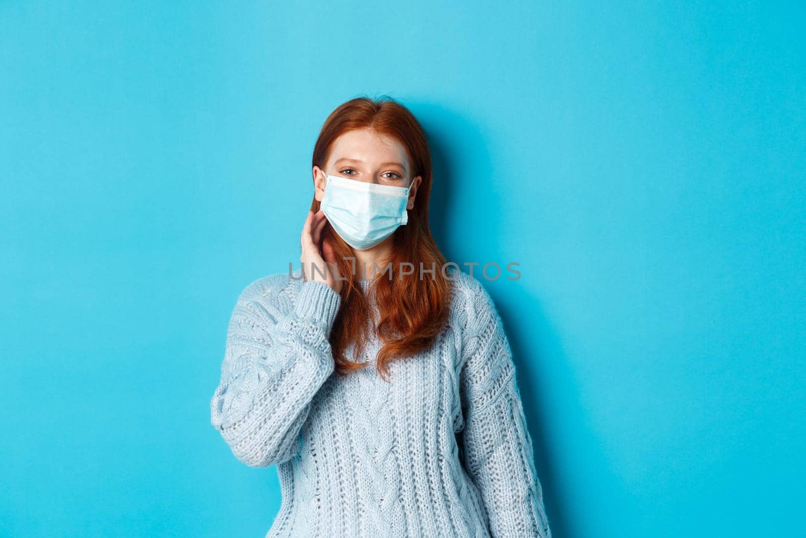 Winter, covid-19 and social distancing concept. Beautiful redhead teenage girl, wearing face mask and tuck hair strand behind ear, staring at camera, standing over blue background.