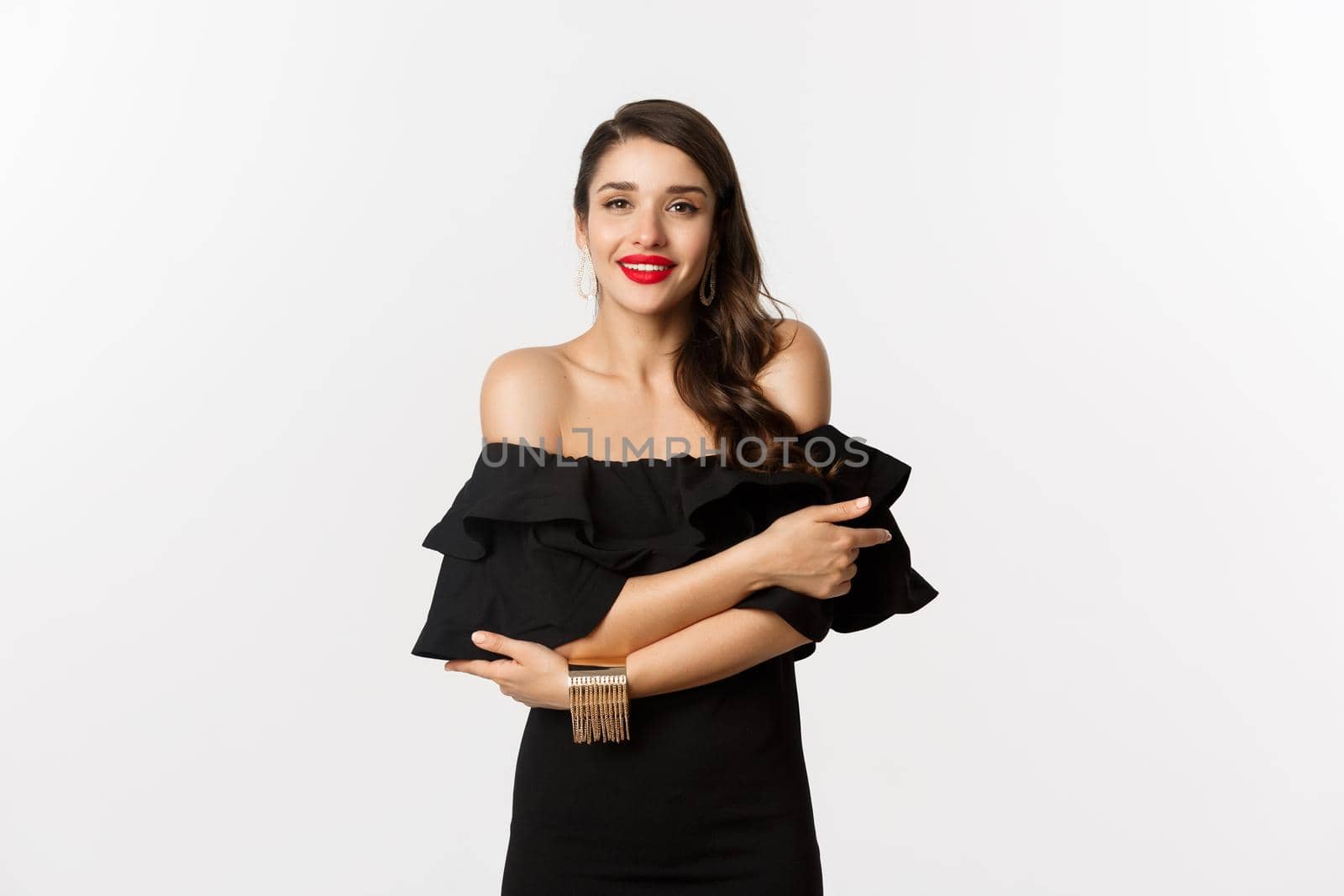 Beauty and fashion concept. Elegant and beautiful woman in black dress, makeup, hugghing herself and looking at camera with sensual gaze, standing over white background.