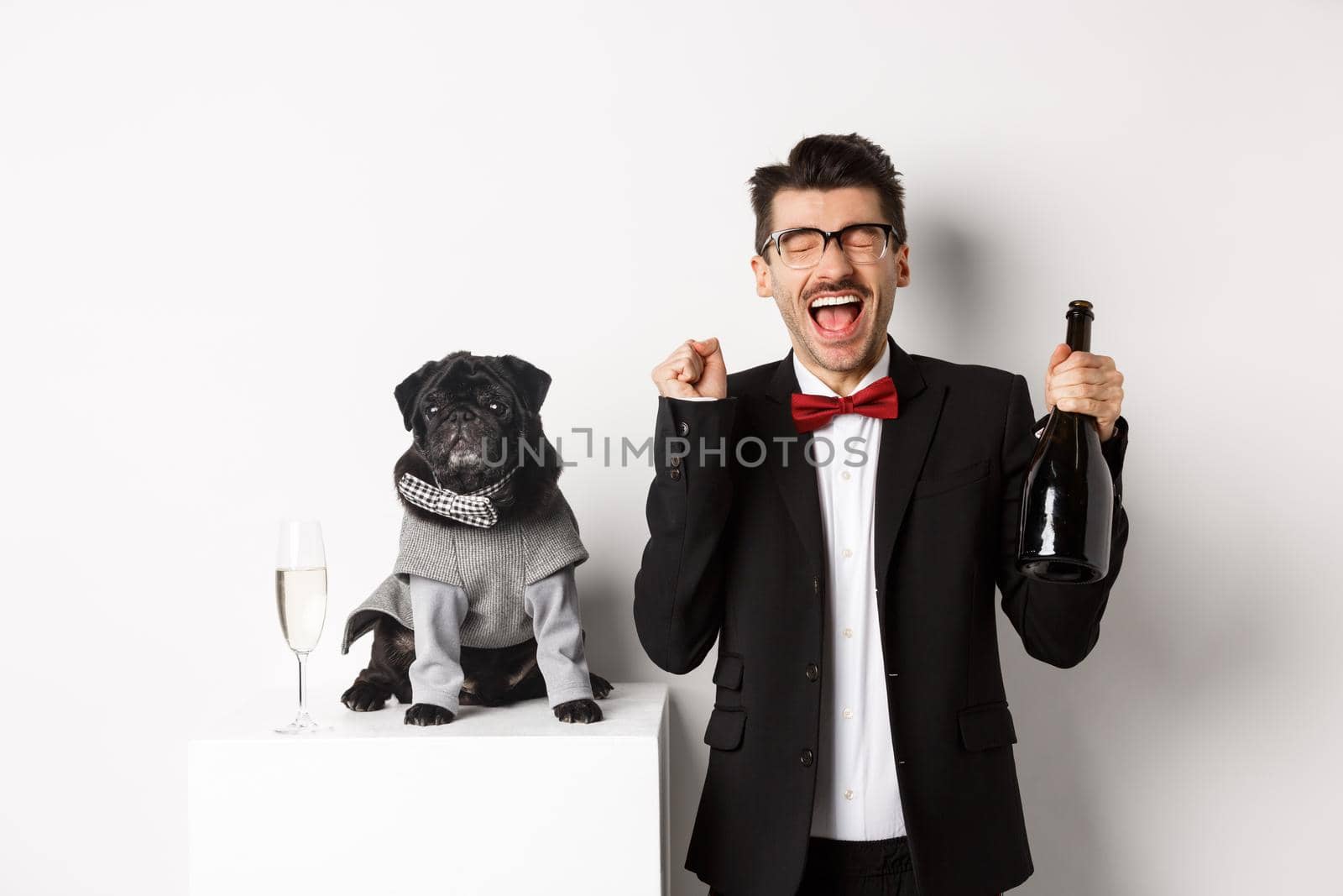 Pets, winter holidays and New Year concept. Happy young man celebrating Christmas with cute black dog wearing party costume, holding bottle champagne, white background by Benzoix