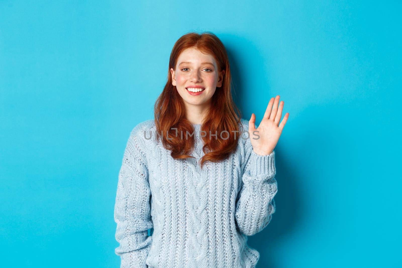 Friendly redhead teenage girl saying hi, waving hand in hello gesture and smiling, standing against blue background by Benzoix