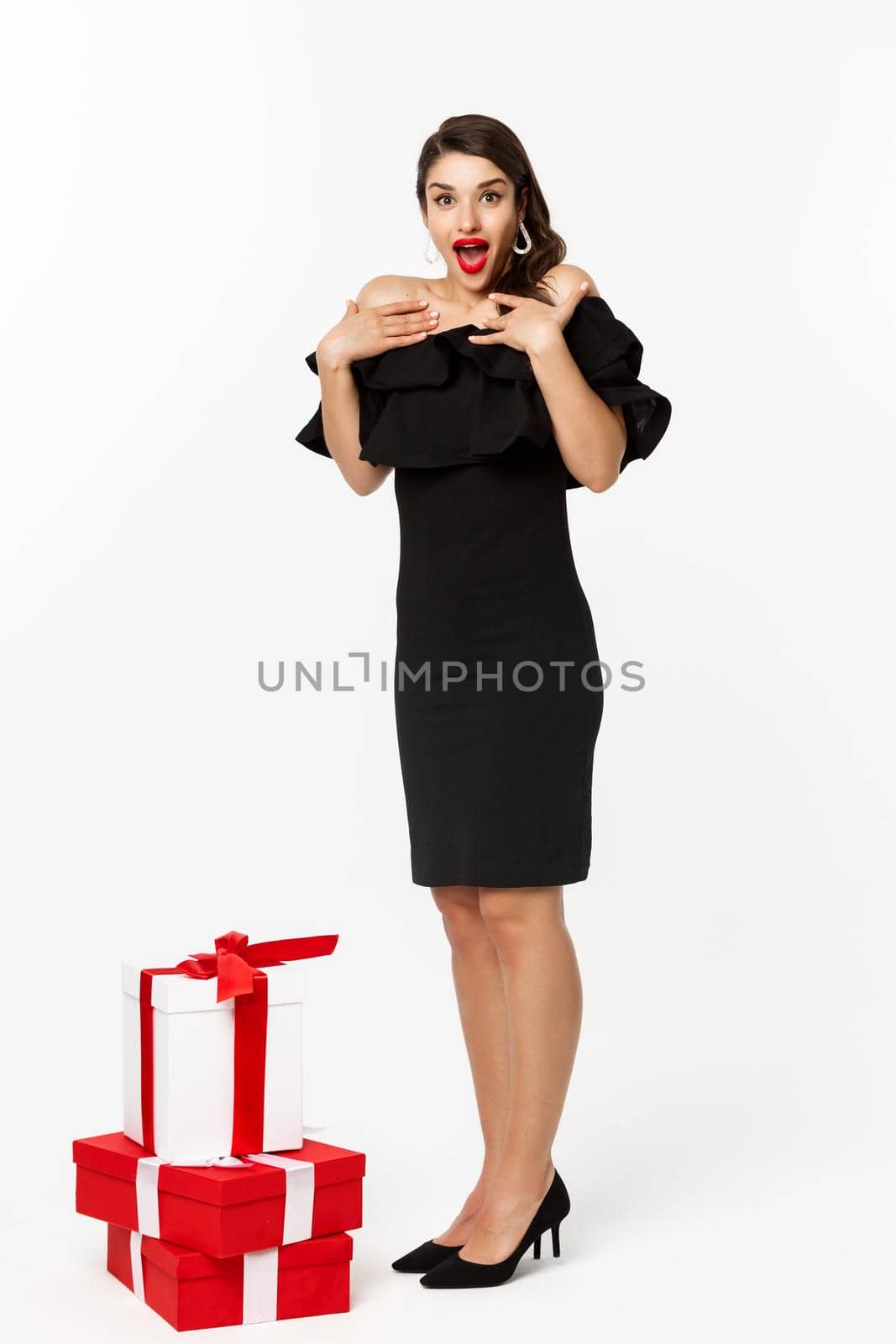 Full length view of woman in elegant dress and red lips, looking surprised, receive gifts on christmas holidays, standing with presents over white background.