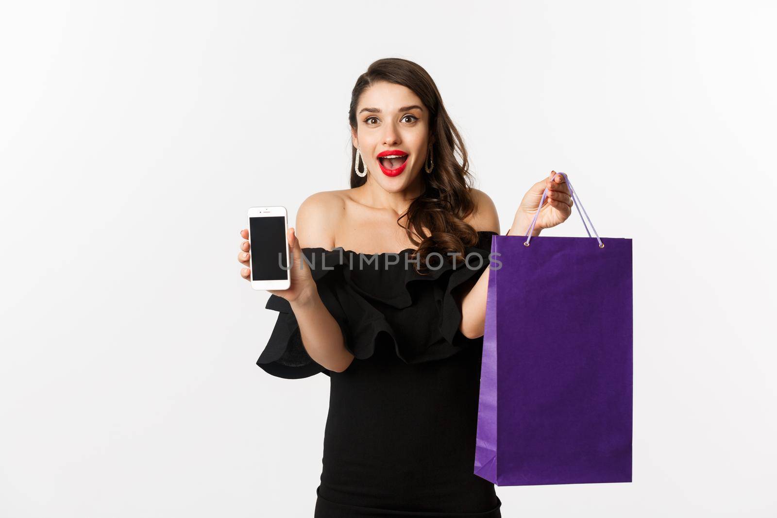 Beauty and shopping concept. Beautiful and stylish woman showing smartphone screen and bag, buying online, standing over white background.