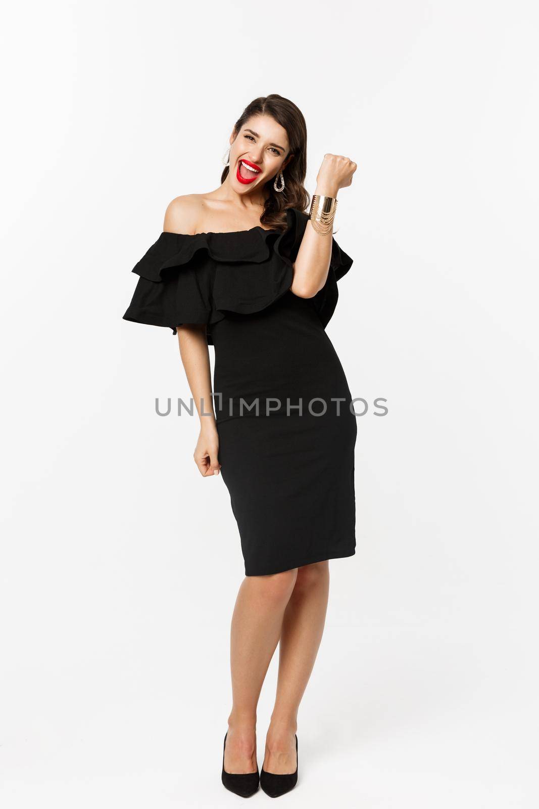 Beauty and fashion concept. Full length shot of happy young woman in luxury black dress, jewelry and heels, wearing makeup and looking satisfied, celebrating victory, winning, white background by Benzoix