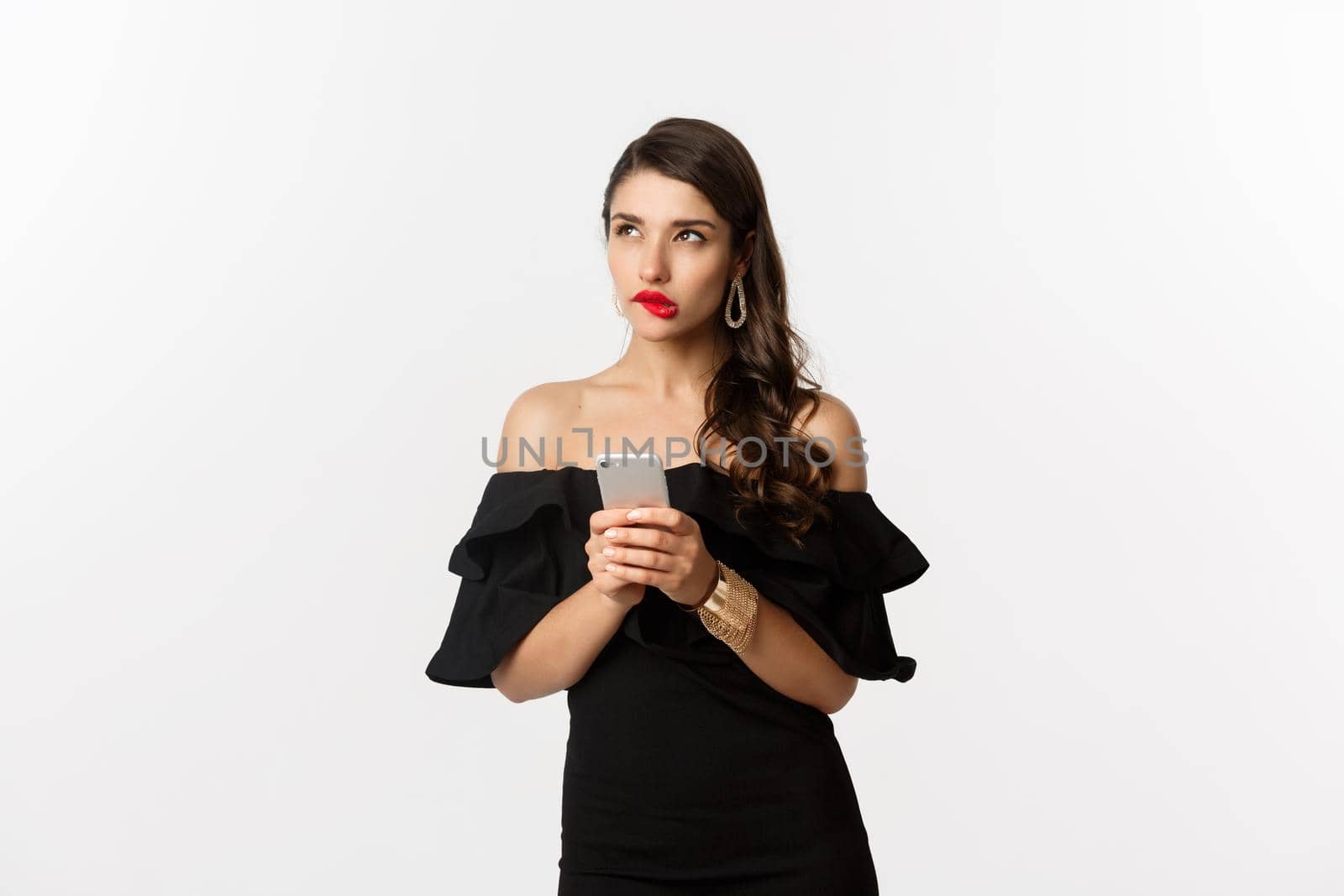 Thoughtful young woman using smartphone and thinking, looking up and pondering, standing over white background. Copy space