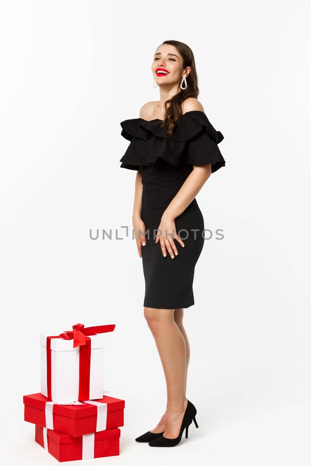 Vertical view of beautiful young woman in luxurious dress, red lips and jewelry, standing with christmas gifts on white background, standing over white background.