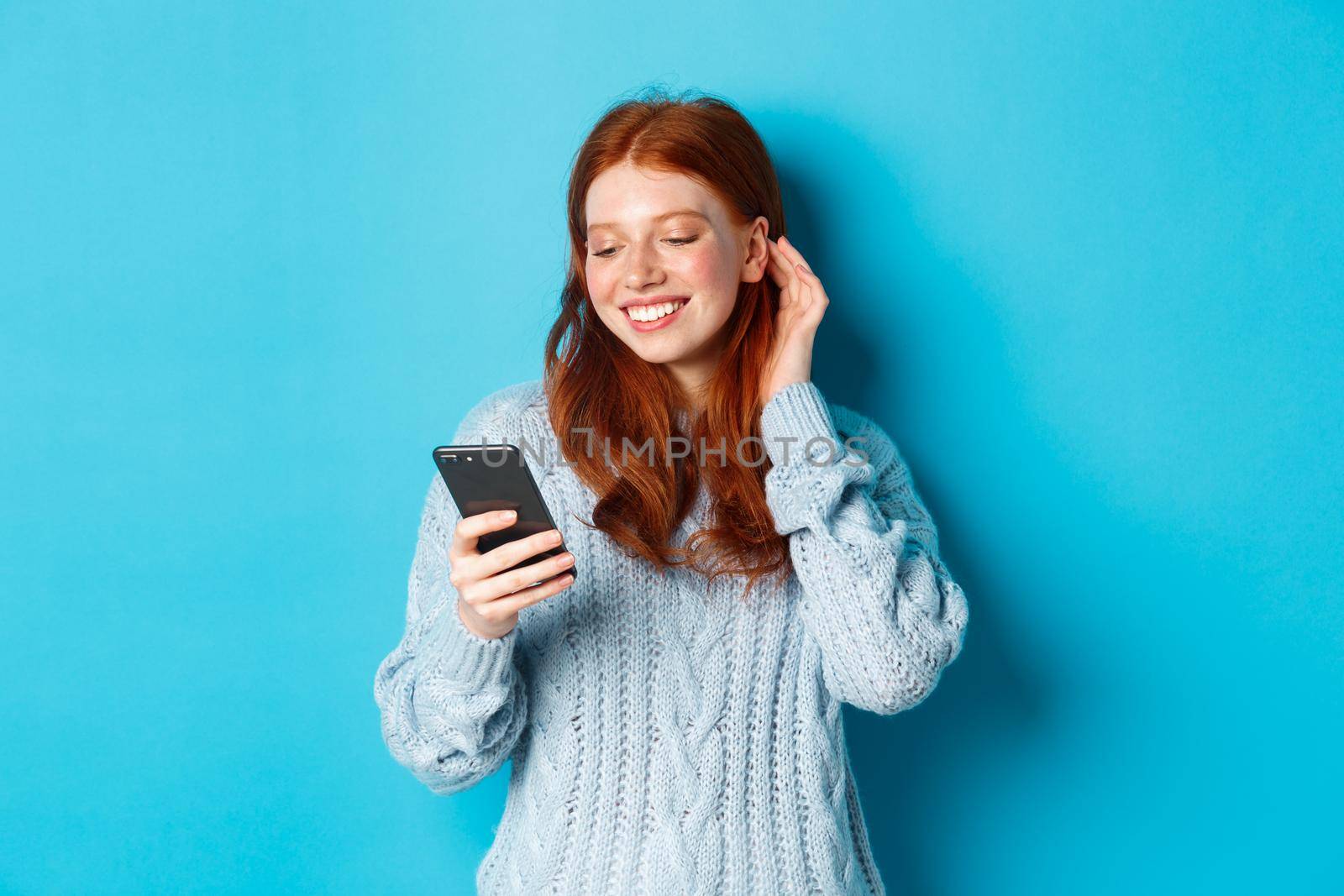 Technology. Happy redhead girl looking pleased at mobile phone, reading compliment in message, smiling and tuck hair behind ear, standing in sweater over blue background.