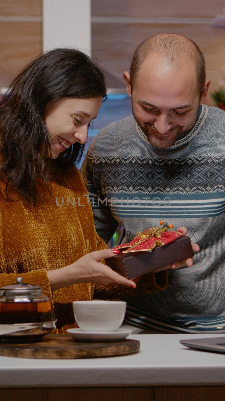 Couple celebrating christmas eve on video call conference with relatives and receiving gifts in holiday decorated kitchen. Man and woman feeling cheerful for seasonal celebration