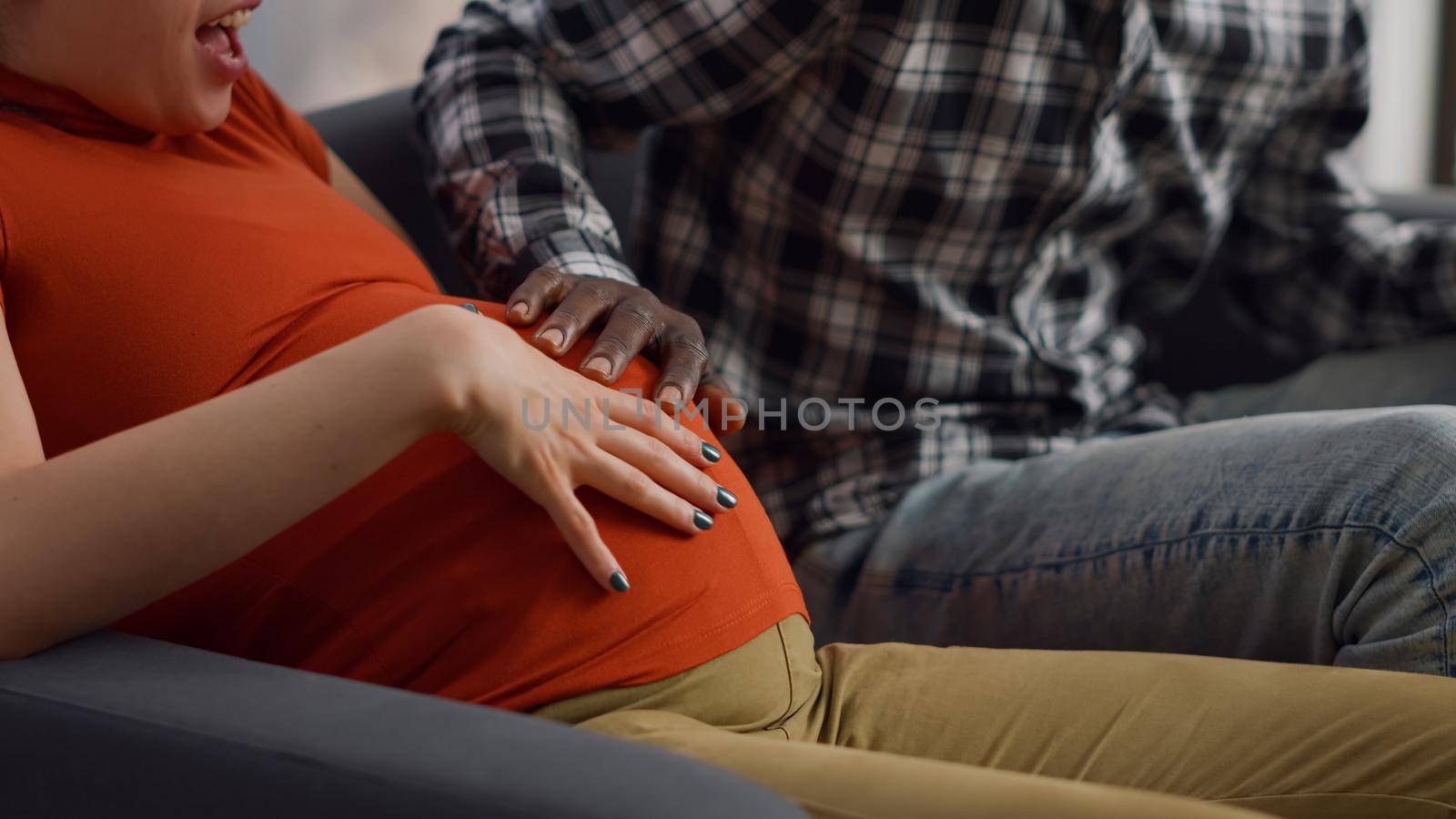 Close up of interracial hands of young couple on belly expecting child while sitting on sofa. Multi ethnic people touching baby bump feeling cheerful and talking about parenthood