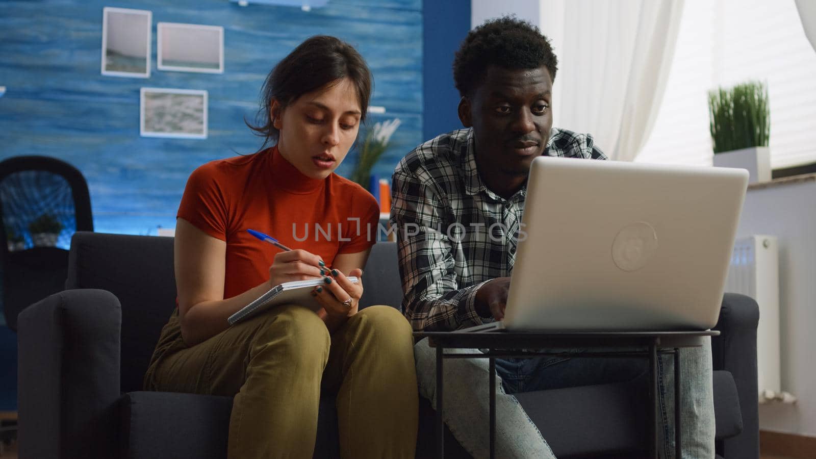 Young interracial couple calculating taxes and paperwork using laptop and notebook in living room. Mixed race husband and wife checking bills and planning budget for tax payment at home