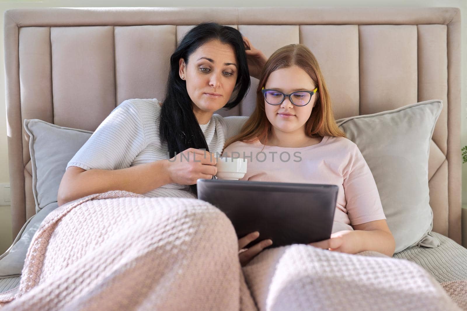 Mother and teenage daughter resting at home together, use digital tablet. Woman and teen girl lying in bed, looking at screen, movie video. Family, parent teenager relationship, vacation, technology