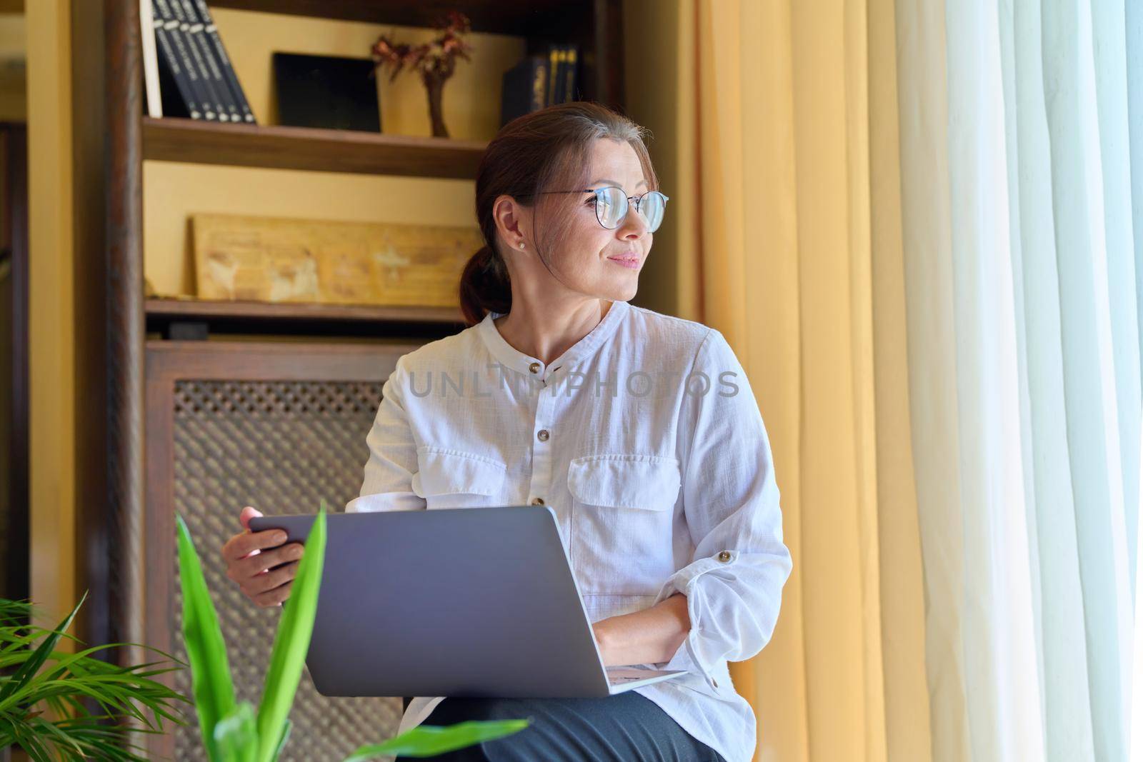 Middle-aged woman psychologist in an office with laptop. Portrait of professional female counselor, psychiatrist, therapist, teacher sitting on chair near window. Technology, medicine, people