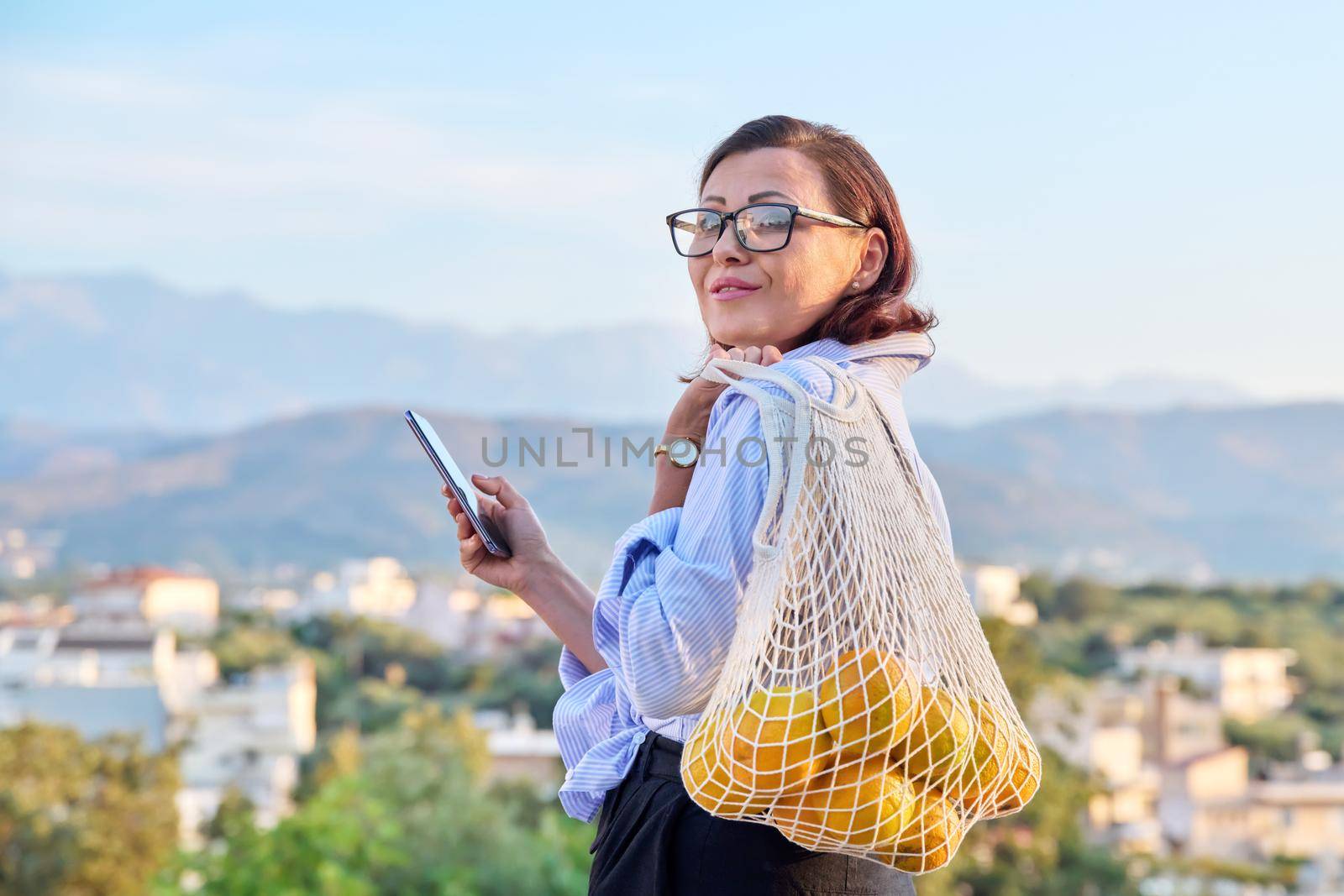 Portrait of beautiful middle-aged woman with smartphone trending grid of oranges outdoors. Female with phone and eco mesh shopping bag with organic oranges, mountains village sunset background, trends