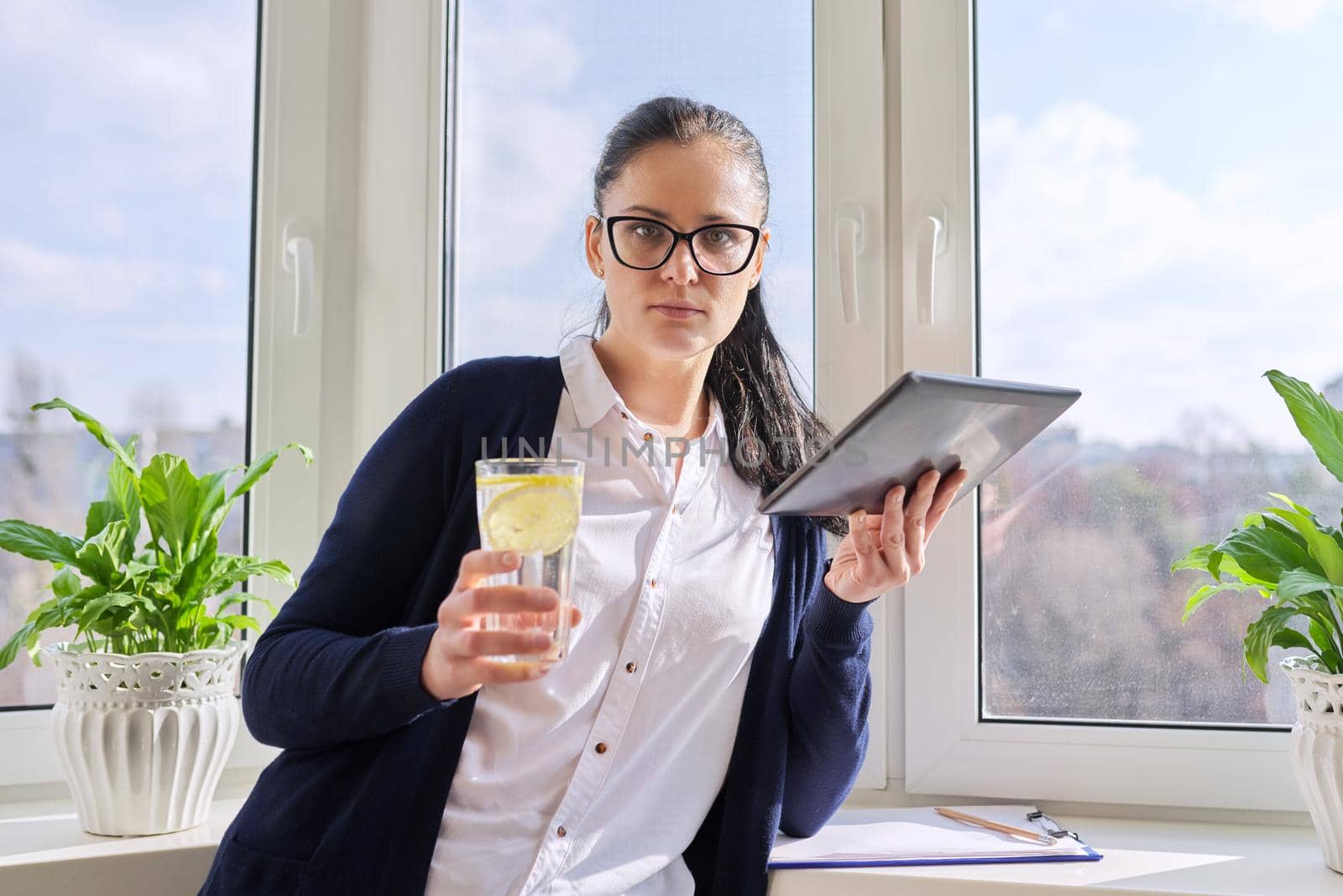 Adult woman drinks water with lemon, female in glasses with digital tablet, at home near window