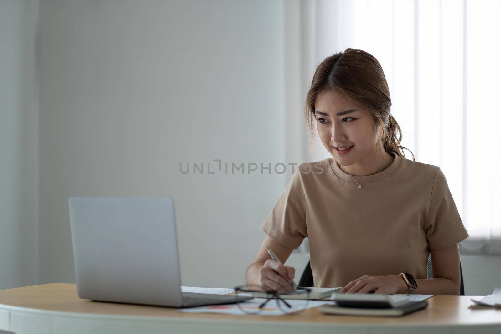 portrait of smiling woman making notes while taking part in webinar at tabletop with laptop in office.