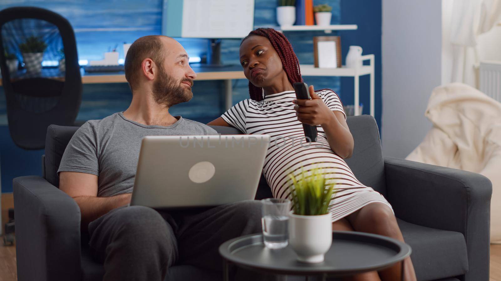 Married interracial couple talking about pregnancy while relaxing together in living room. Multi ethnic people using laptop and watching television. Black woman holding TV remote control