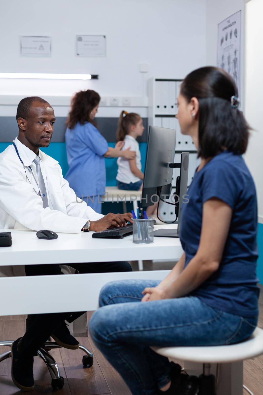 African american pediatrician doctor explaining sickness expertise to patient mother typing medical treatment on computer. Specialist man working in hospital office during clinical appointment