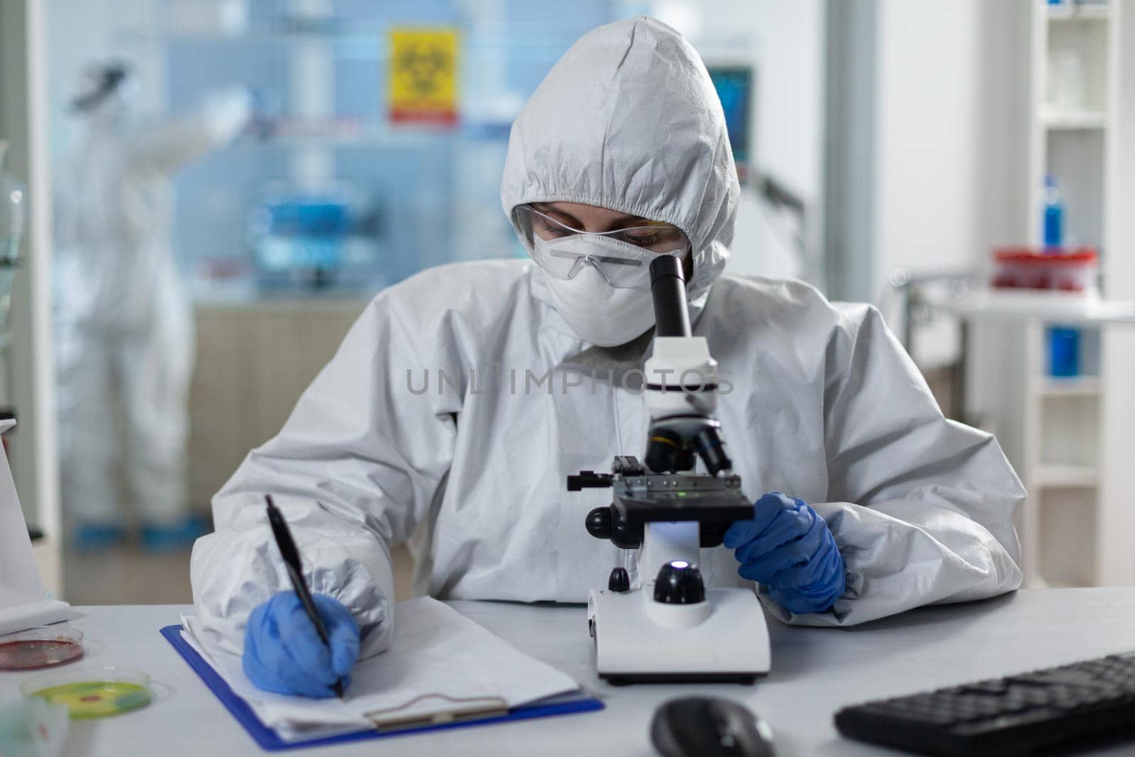 Biologist researcher wearing protective suit analyzing chemicals using medical microscope while writing clinical expertise during scientific experiment. Specialist doctor working in biochemistry lab