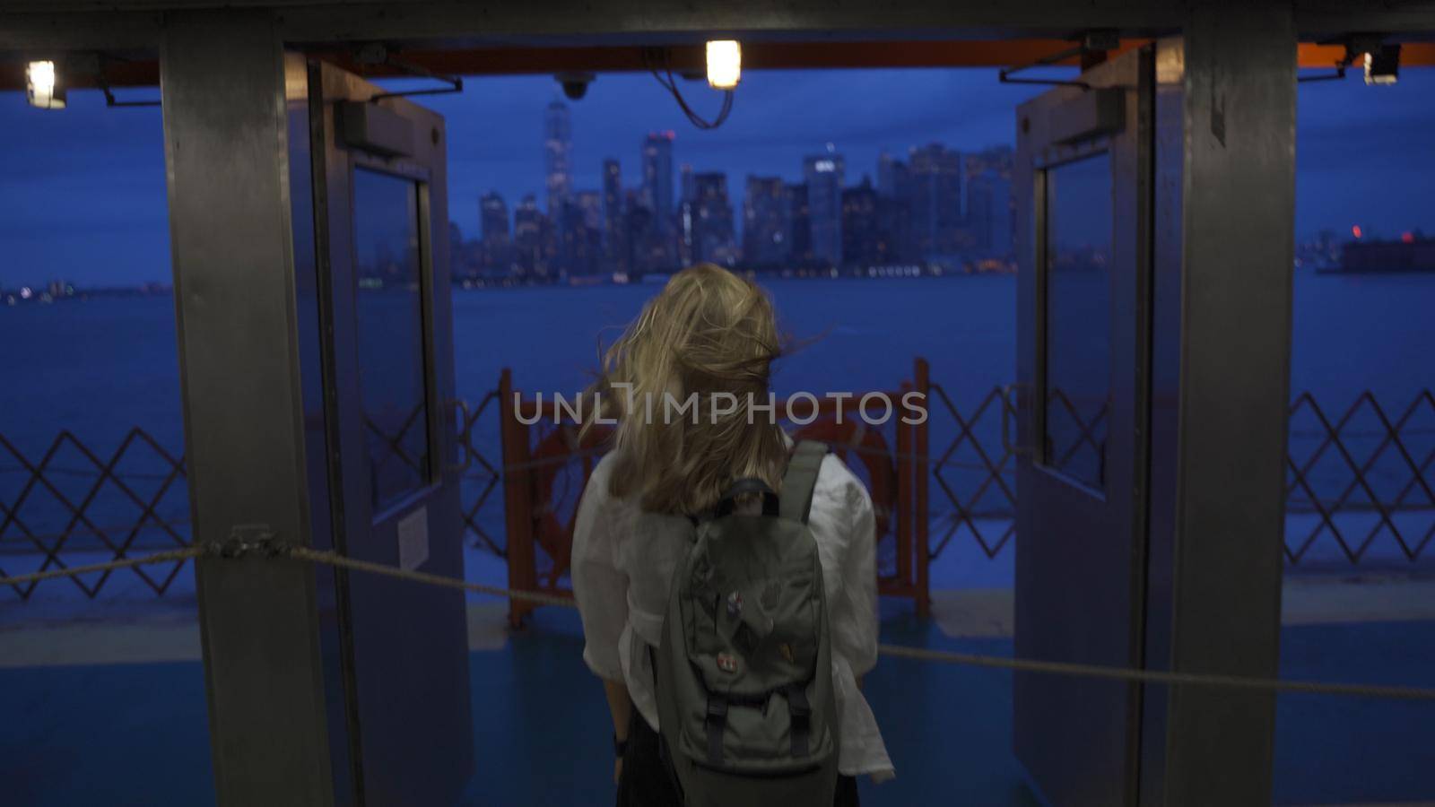 A female looks off into the distance, deep in thought while staring at the New York City skyline while riding the Staten Island Ferry. Back view of tourist on Staten Island Ferry approaching Manhattan in the evening.