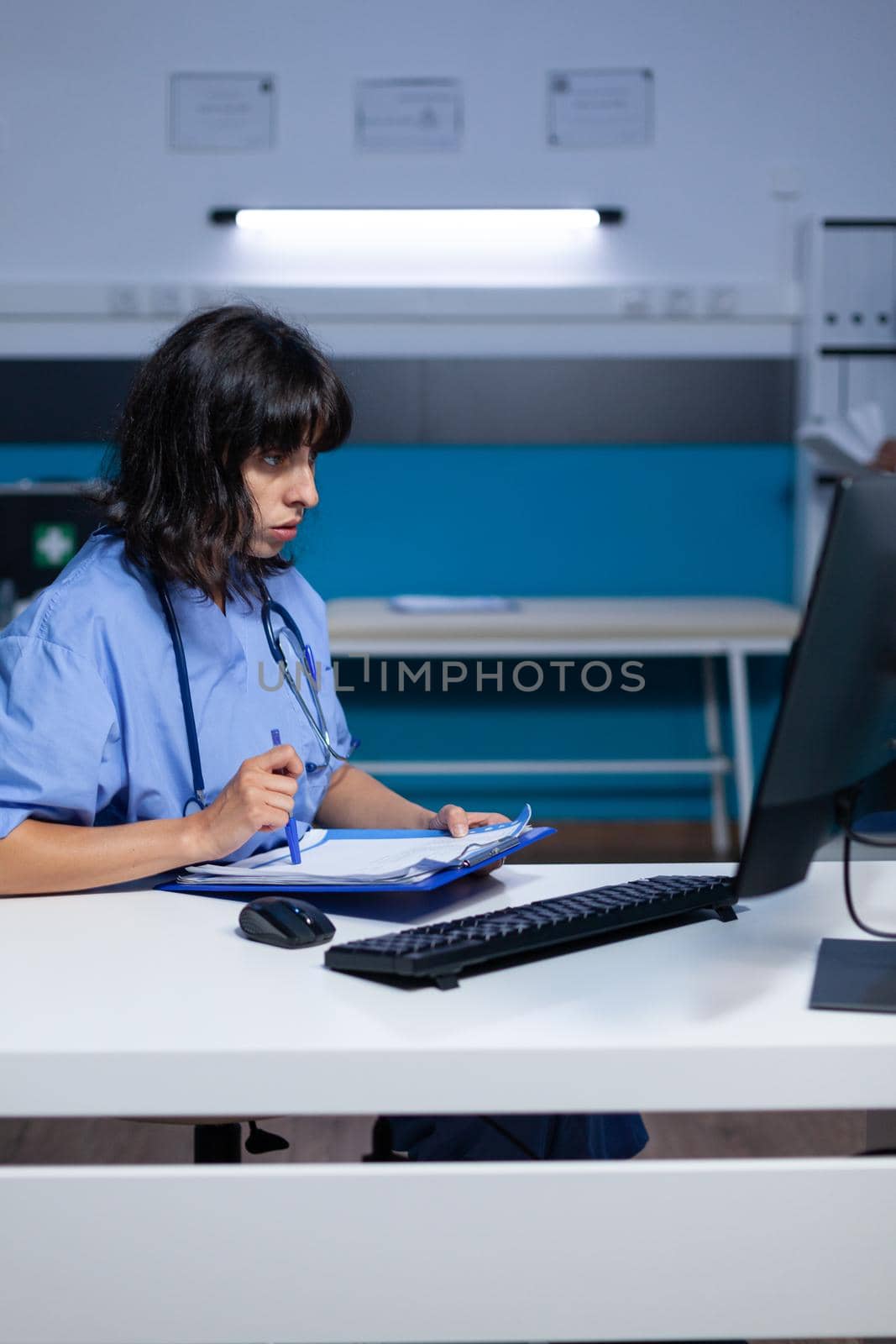 Medical assistant analyzing documents and files on monitor, working late at night. Woman nurse looking at computer and working on healthcare appointment with checkup papers for patient.