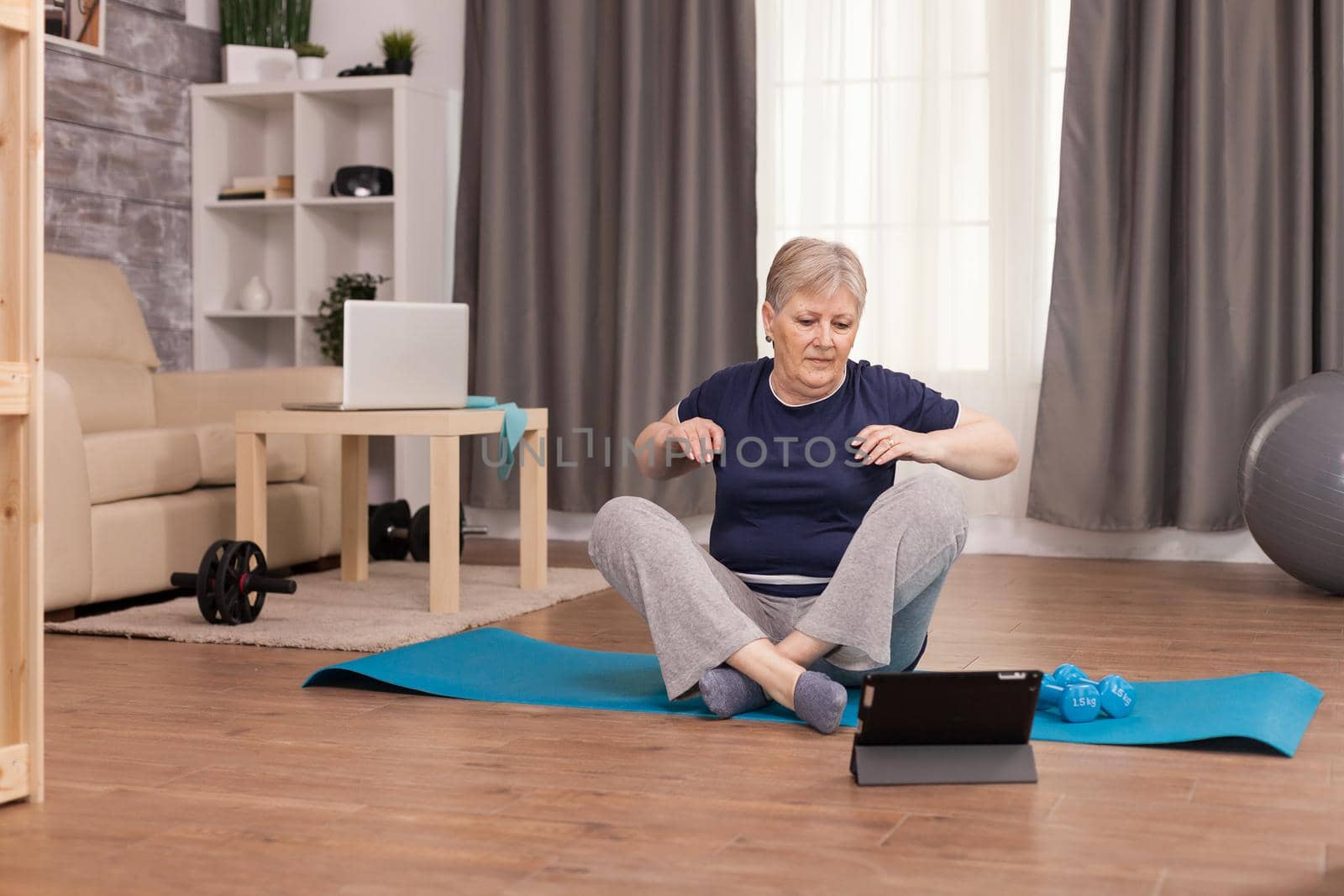 Retired woman warming up her shoulders before starting the fitness training. Old person pensioner online internet exercise training at home sport activity with dumbbell, resistance band