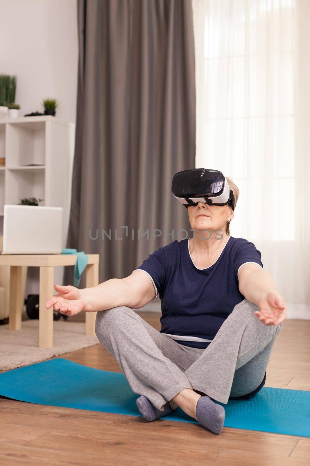 Senior woman using VR headset during yoga exercise. Old person pensioner online internet exercise training at home sport activity with dumbbell, resistance band, swiss ball and VR headset at elderly retirement age