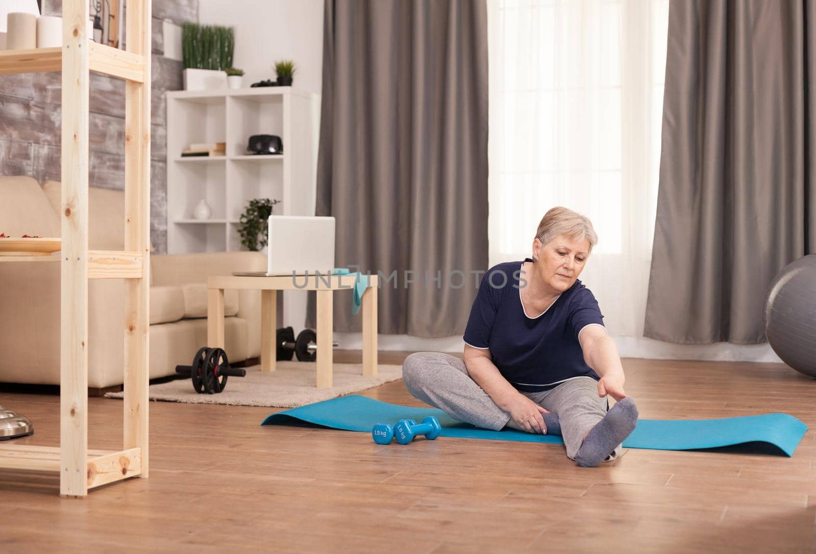 Pensioner woman stretching at home on the yoga mat. Old person pensioner online internet exercise training at home sport activity with dumbbell, resistance band, swiss ball at elderly retirement age