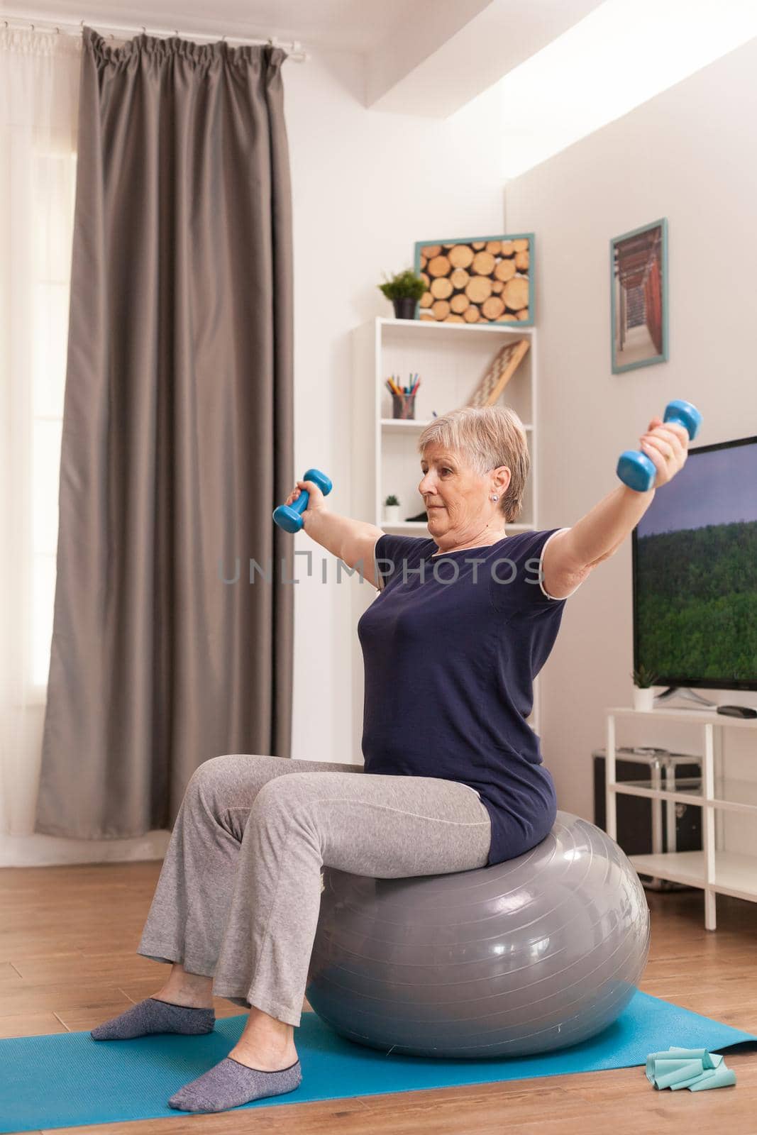 Senior woman using balance ball and dumbbells sitting on yoga mat at home. Old person pensioner online internet exercise training at home sport activity with resistance band, swiss ball at elderly retirement age
