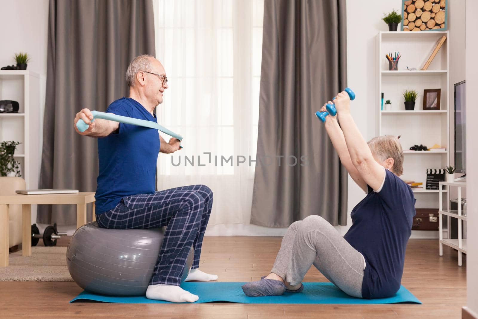 Pensioners doing sport in pajamas with resistance band and dumbbells. Old person healthy lifestyle exercise at home, workout and training, sport activity at home on yoga mat.