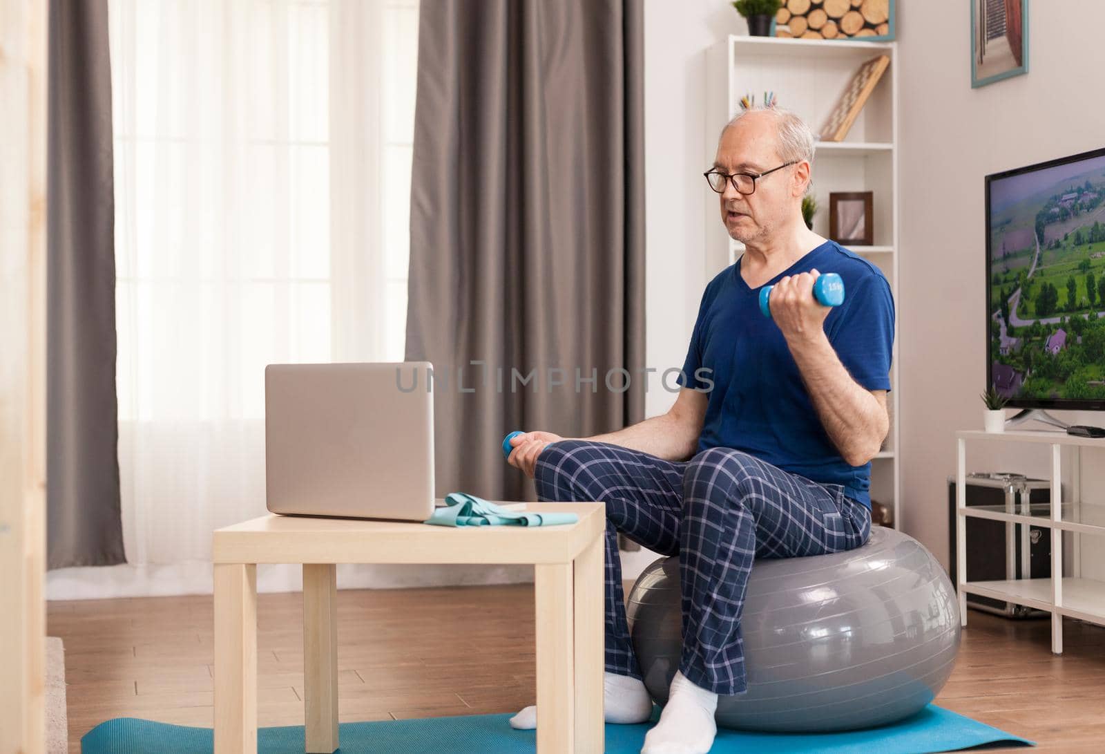 Senior man training with online coach sitting on swiss ball in the middle of the room. Old person pensioner online internet exercise training at home sport activity with dumbbell, resistance band, swiss ball at elderly retirement age.