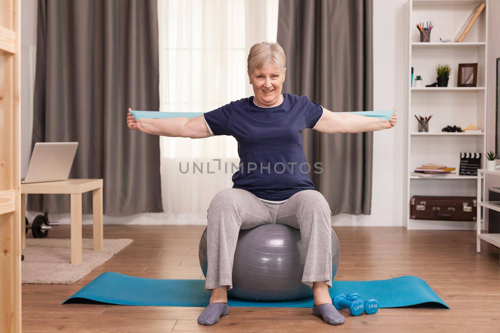 Senior woman working out at home with fitness bands to solve her health problems. Old person pensioner online internet exercise training at home sport activity with dumbbell, resistance band, swiss ball at elderly retirement age